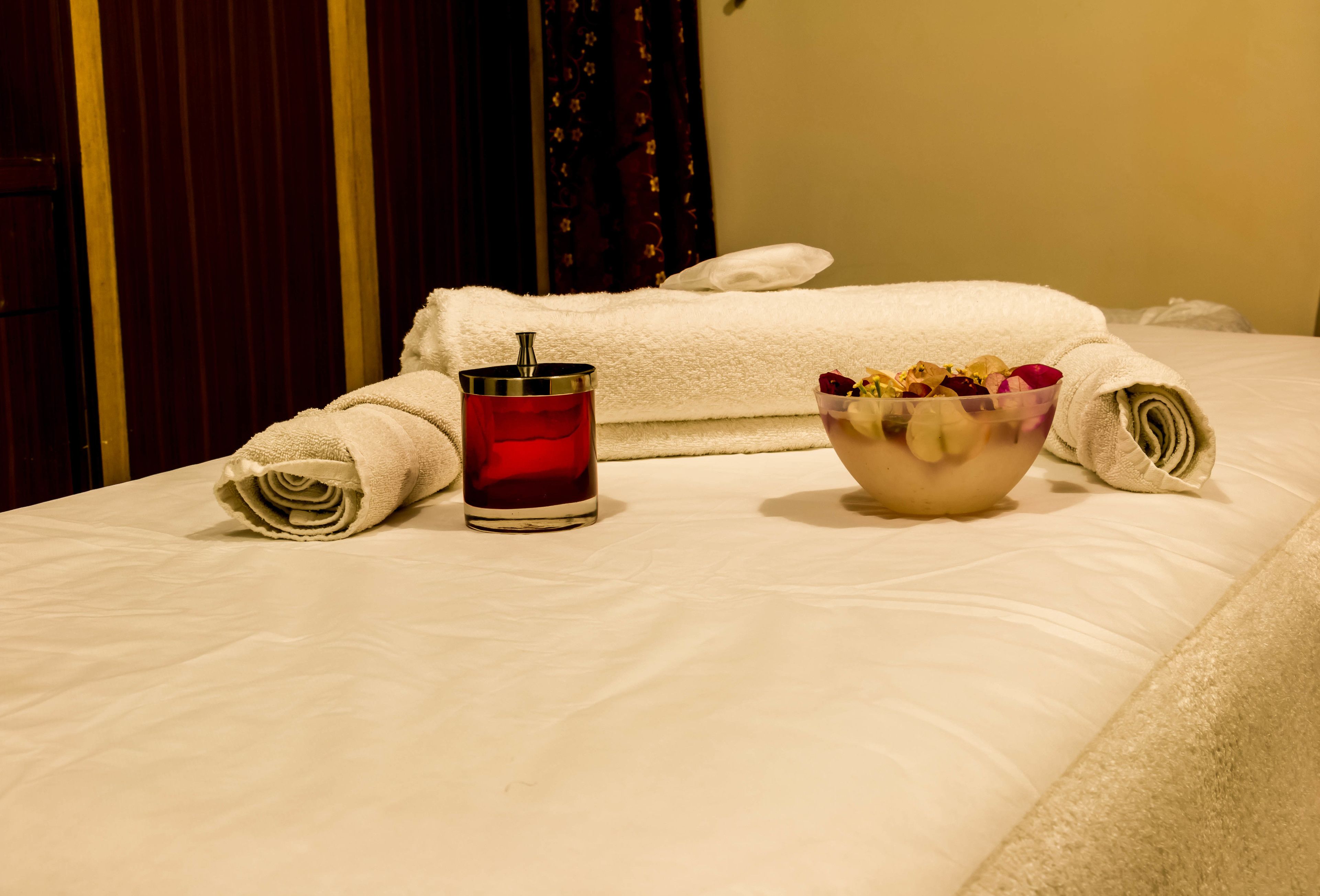 A neatly made bed with a rolled towel, a glass of red beverage, and a bowl of fruit on a hotel room bed.