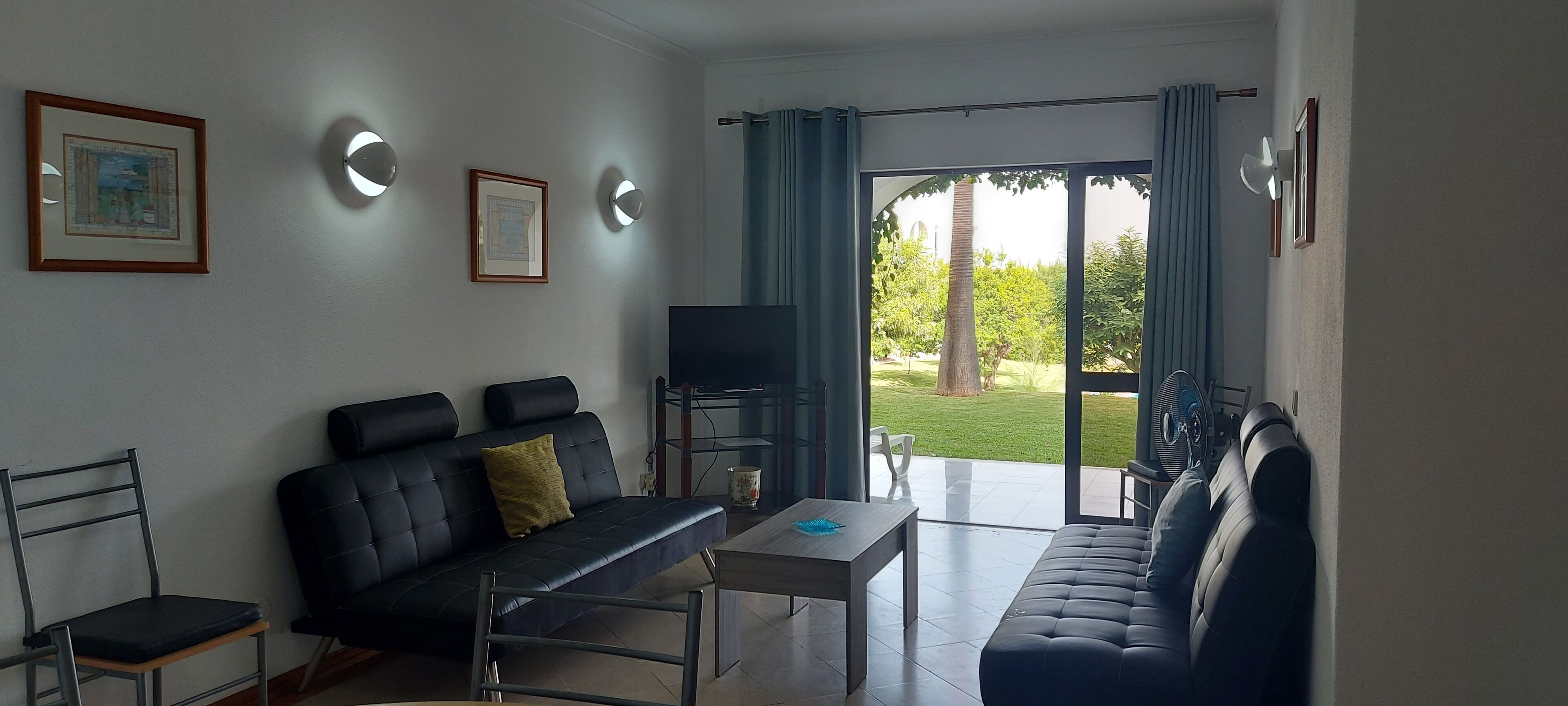 A living room with a sofa, a TV, and a table. There is a large window with curtains.