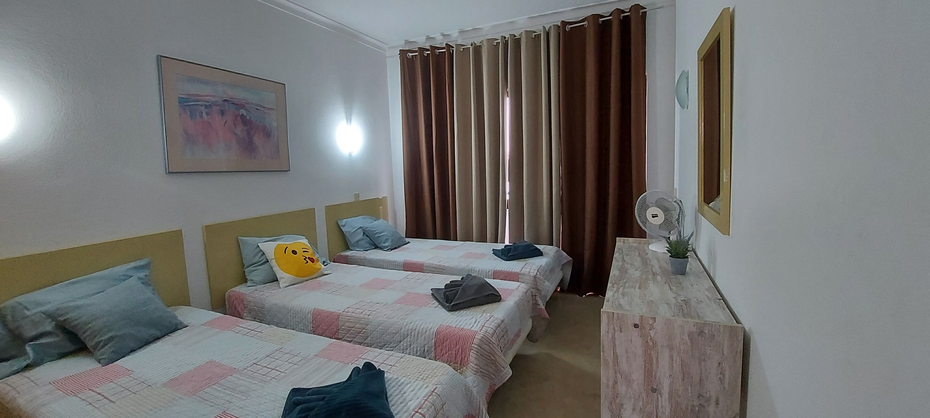 A hotel room with two beds, a bedside table with a fan, a painting on the wall, and a window with curtains.