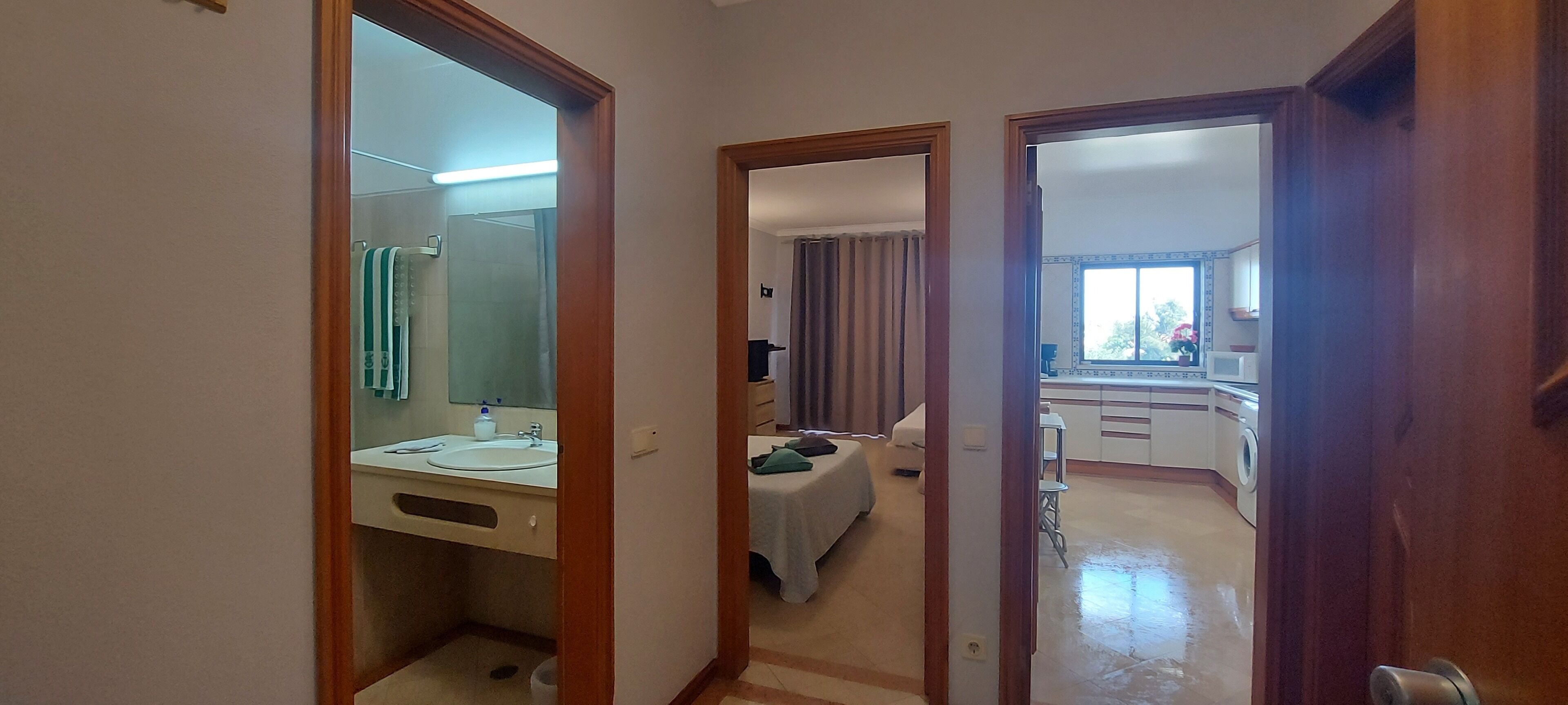 A hallway leading to a bathroom with a sink and mirror, a bedroom with a bed and curtains, and a kitchen area with a window.