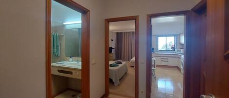 A hallway leading to a bathroom with a sink and mirror, a bedroom with a bed and curtains, and a kitchen area with a window.