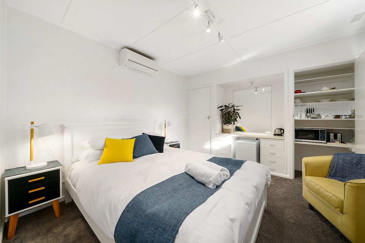 Habitación de hotel moderna con una cama bien hecha, escritorio, silla y zona de cocina integrada.