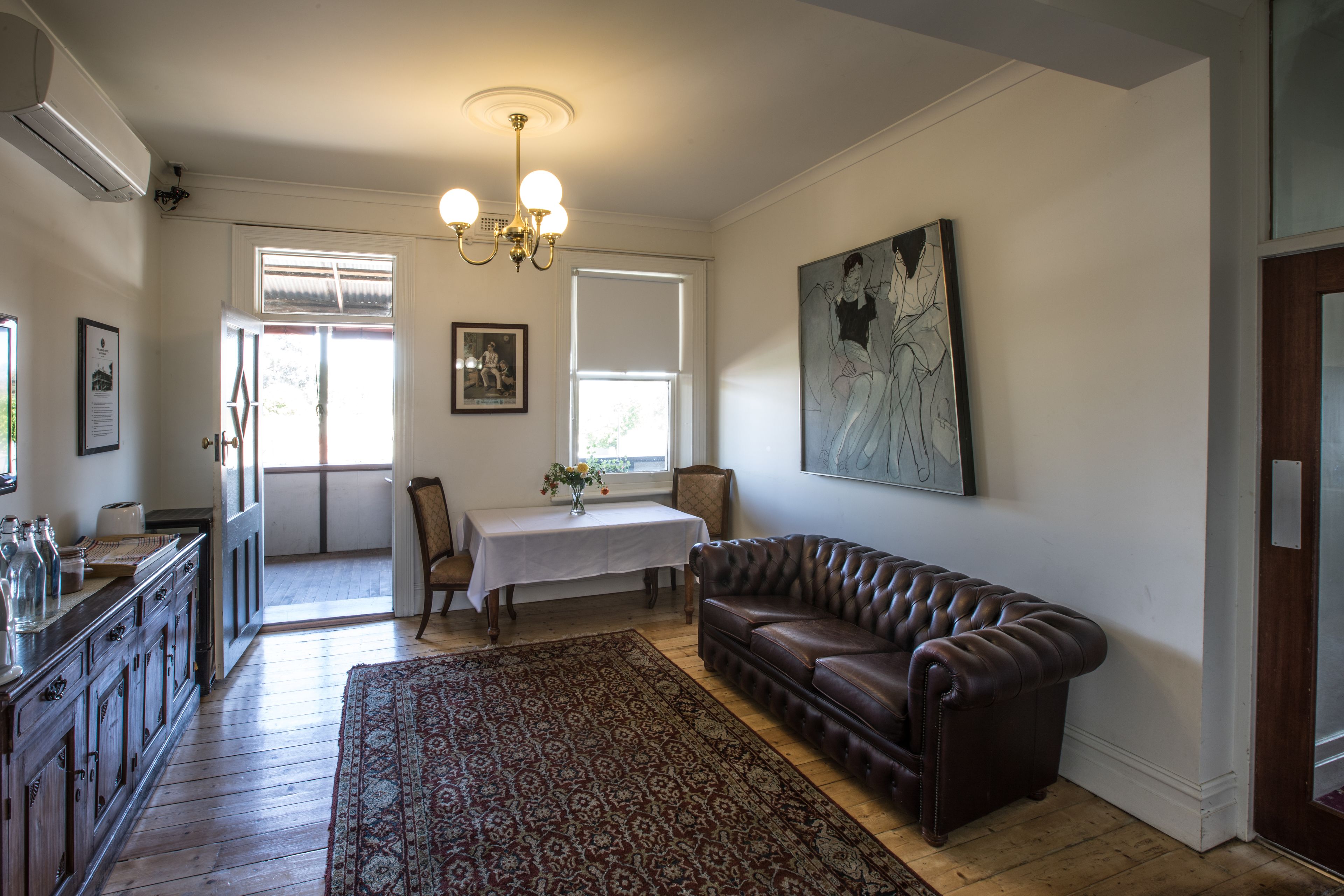 A living room with a brown leather sofa, a patterned rug, a white table with a vase, and a painting on the wall.