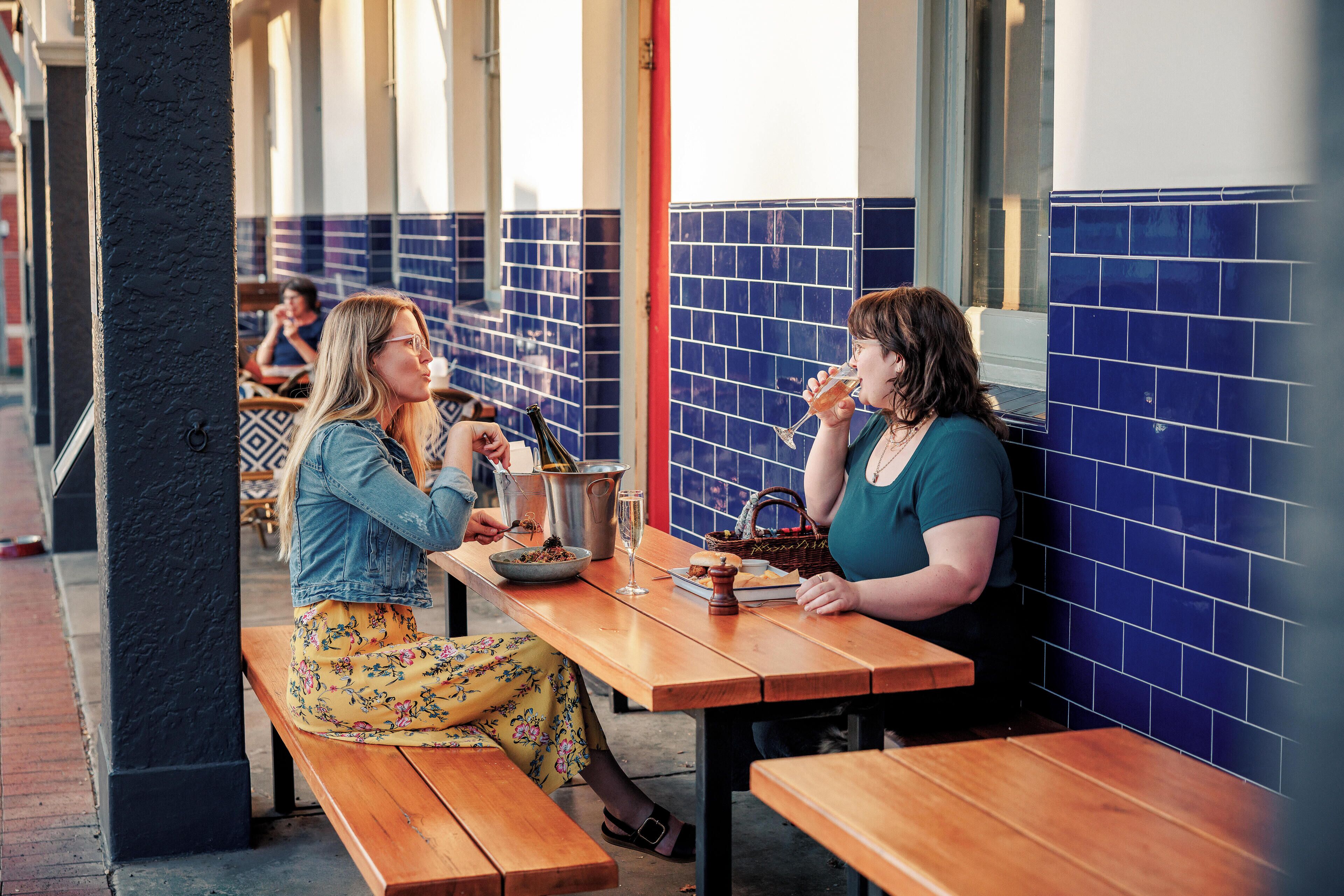 Two people sitting at a wooden table with a tiled wall in the background.