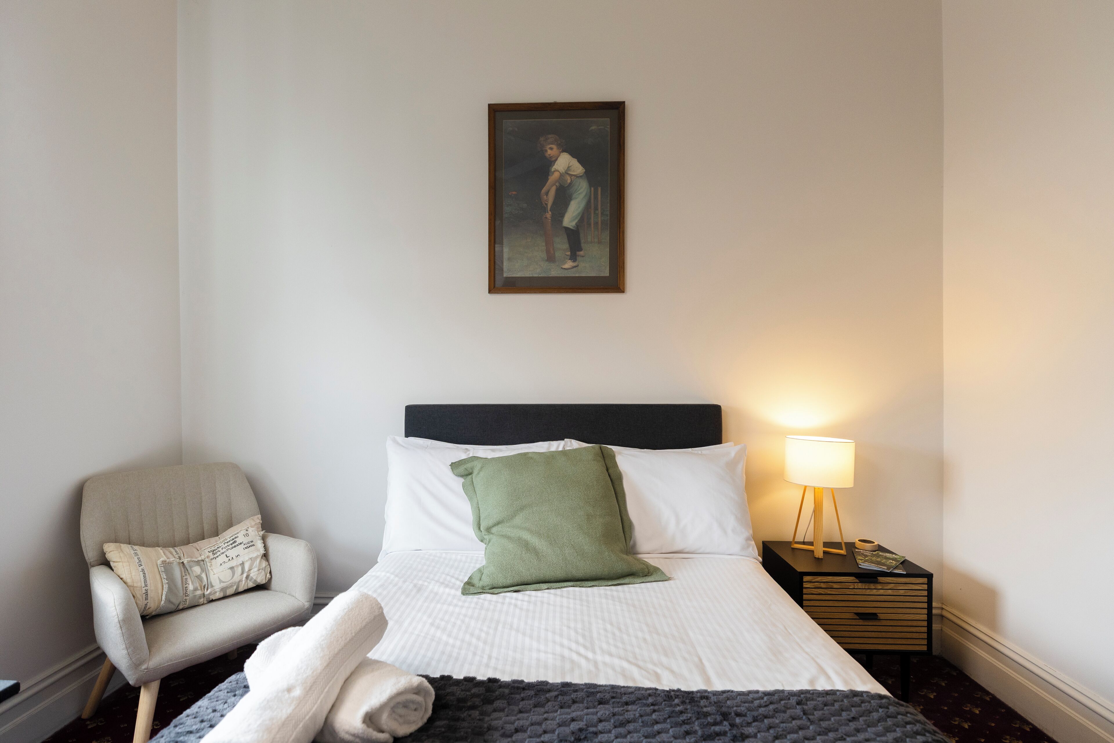 A hotel room with a bed, a chair, a nightstand, and a painting on the wall.