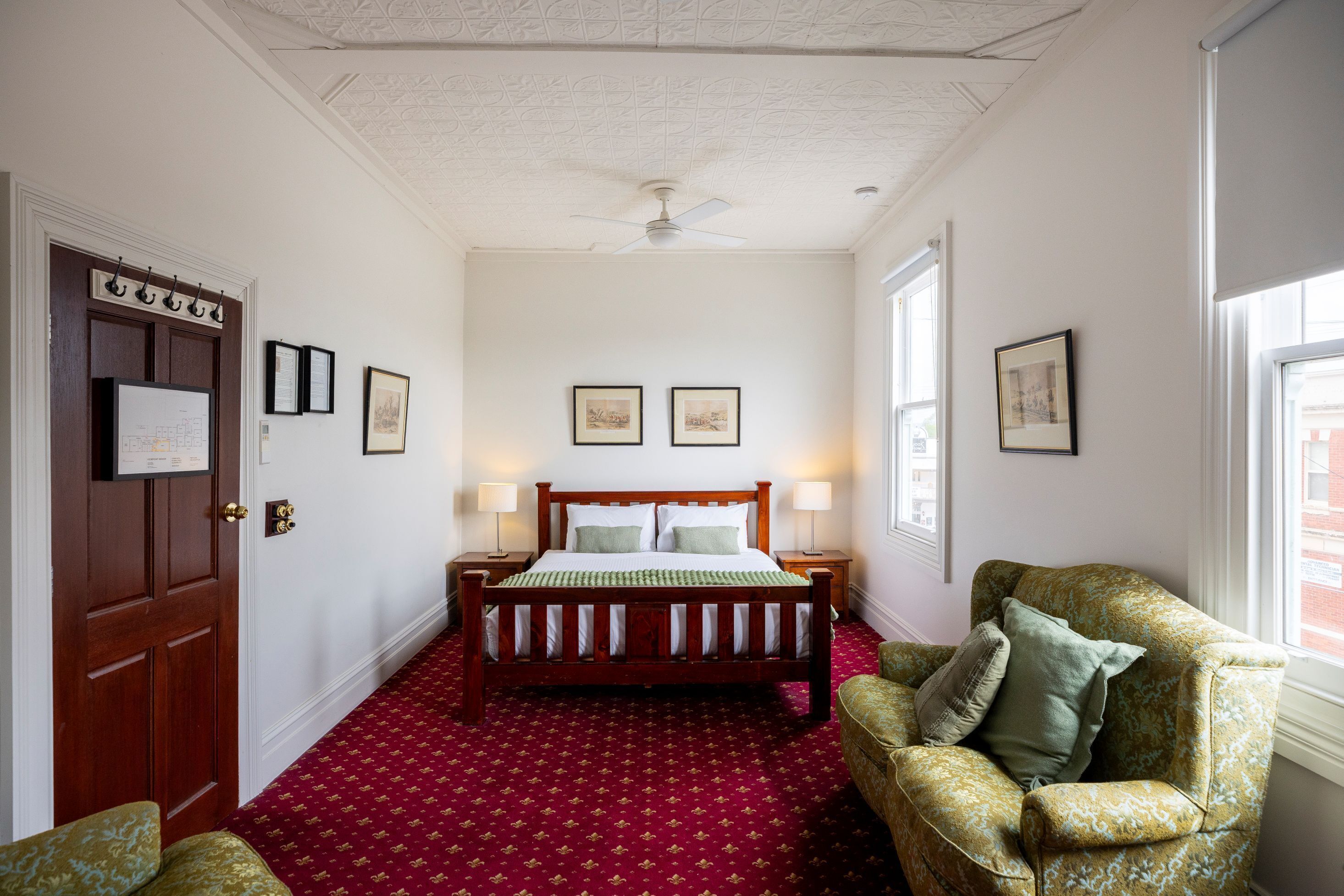 A bedroom with a wooden bed, two bedside lamps, a red patterned carpet, a green armchair, and a door with a coat rack.