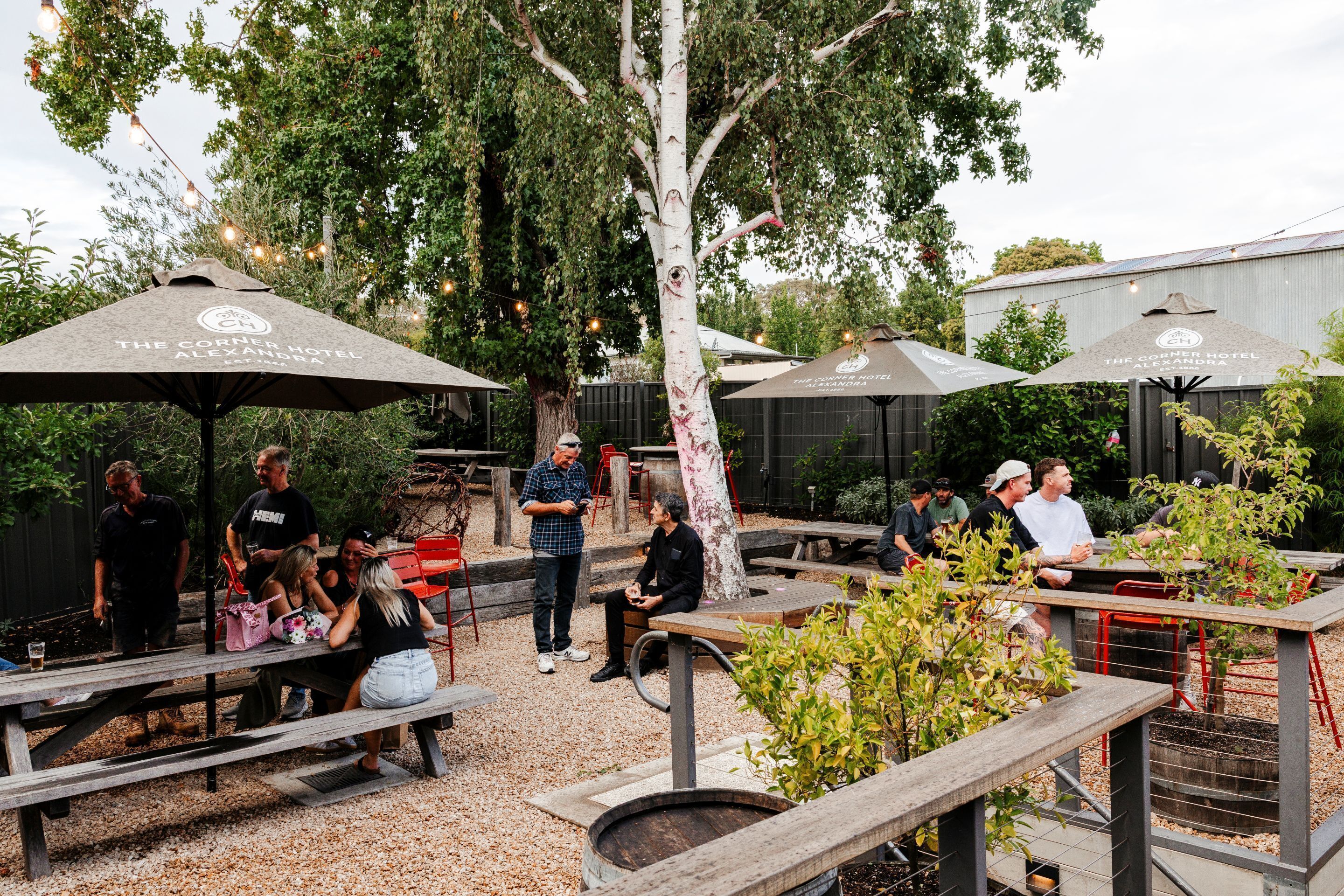 An outdoor dining area with wooden tables and benches, surrounded by trees and covered by umbrellas.