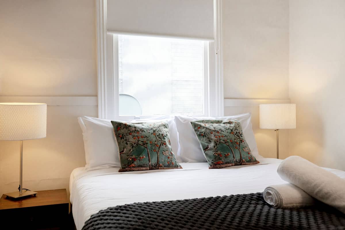 Une chambre d’hôtel avec un lit, deux oreillers à motifs floraux, une lampe de chevet, une fenêtre et une serviette enroulée posée sur le lit.