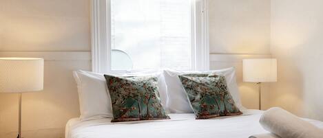 Une chambre d’hôtel avec un lit, deux oreillers à motifs floraux, une lampe de chevet, une fenêtre et une serviette enroulée posée sur le lit.