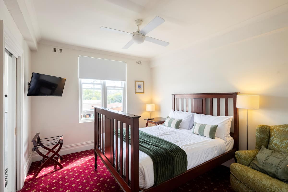Une chambre à coucher avec un lit, un ventilateur de plafond, un téléviseur fixé au mur, une fenêtre avec des stores, une lampe de chevet et un fauteuil vert.