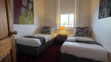 A room with two single beds, a wooden door, a small bedside table with a lamp, and a colorful abstract painting on the wall.