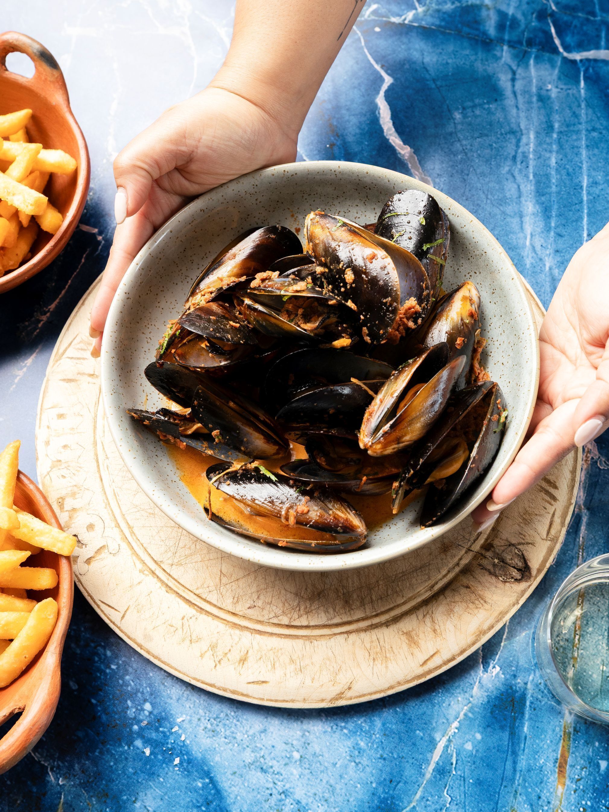 A bowl of mussels on a plate, a wooden board with fries, and a blue textured surface.
