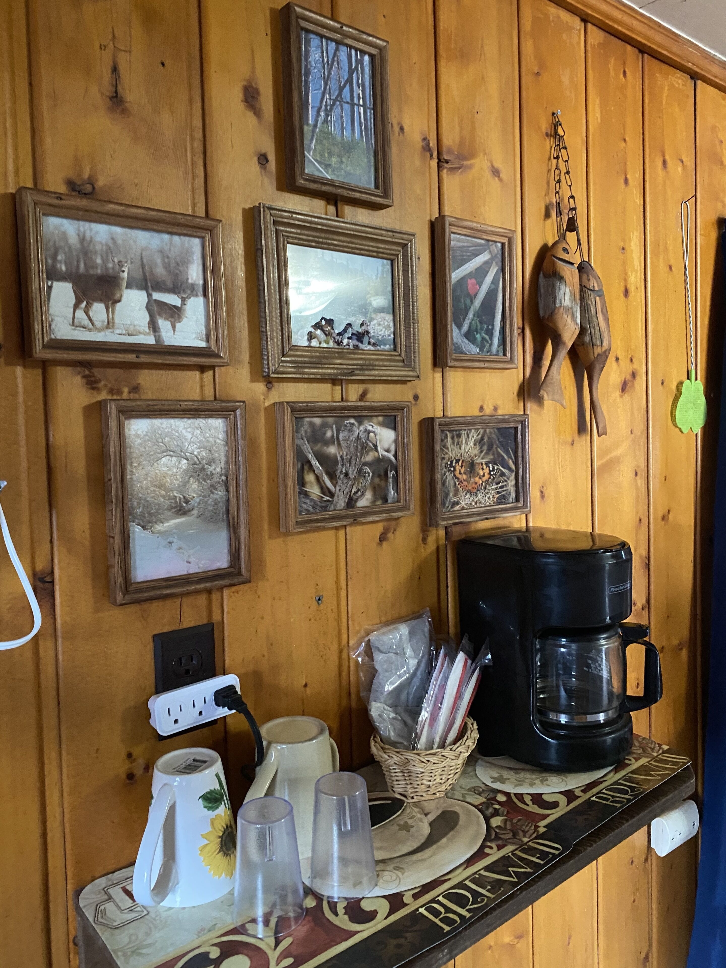 Una pared de madera con cuadros enmarcados, una cafetera y una repisa con tazas y vasos.