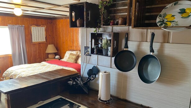 A cozy room with a bed, a small table, a wall-mounted stove, and a shelf with kitchen utensils. - Roundtop Mountain Vista - Cabins and Motel (Thermopolis)