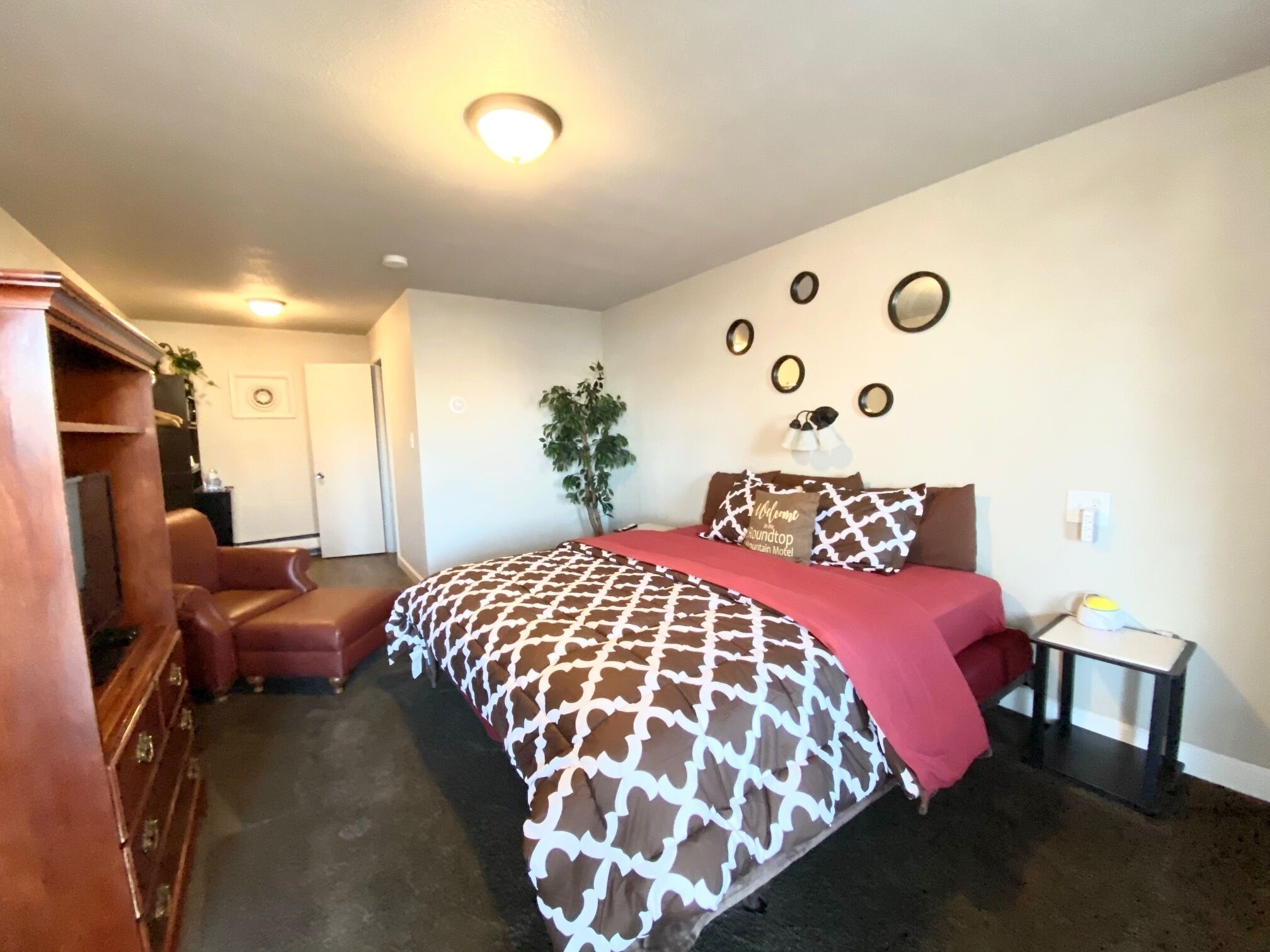 A bedroom with a bed, a dresser, a plant, and a small table with a lamp.