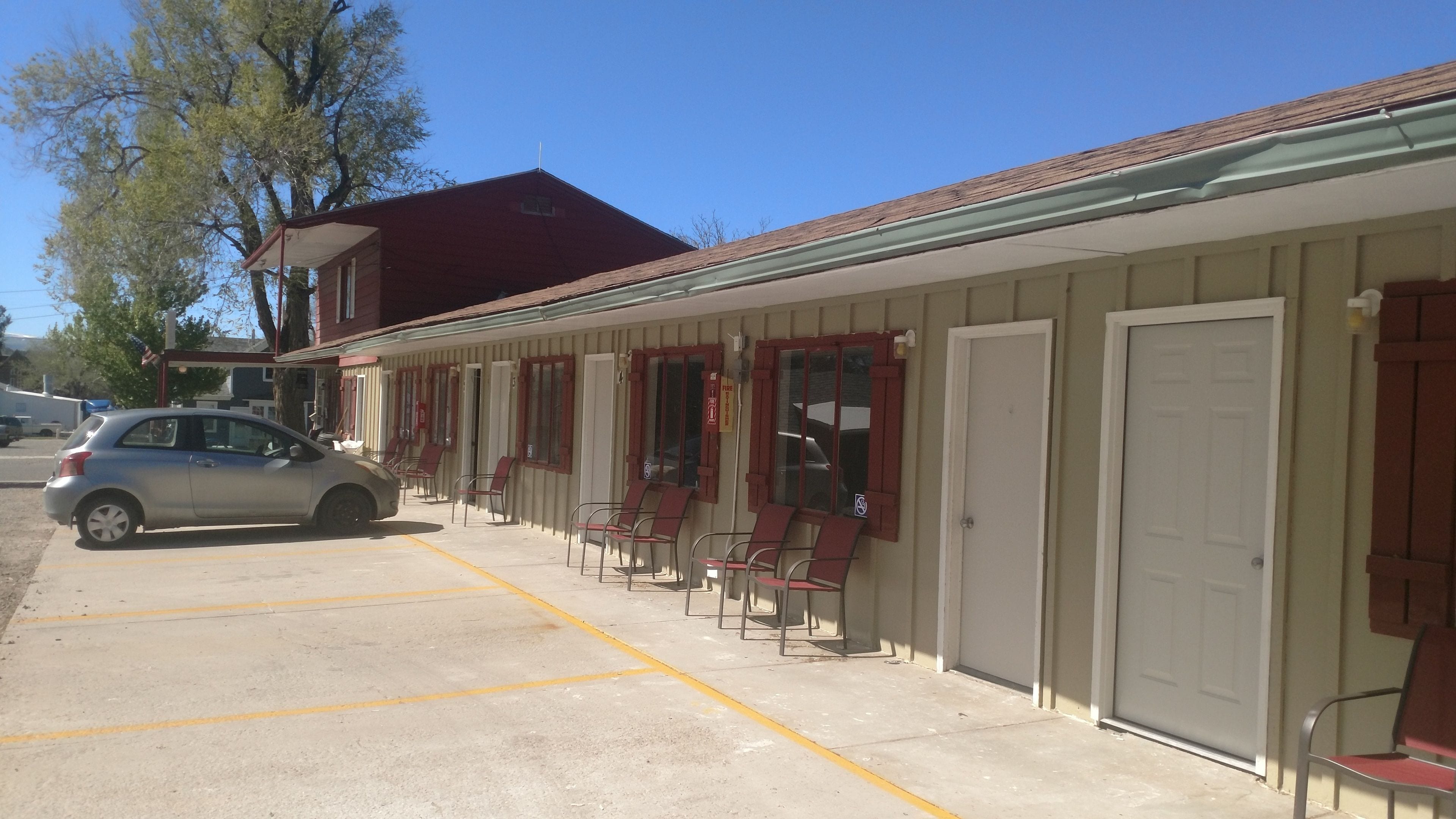 A motel with a row of rooms, each with a door and a window. There are chairs outside the rooms, and a car is parked in the lot.