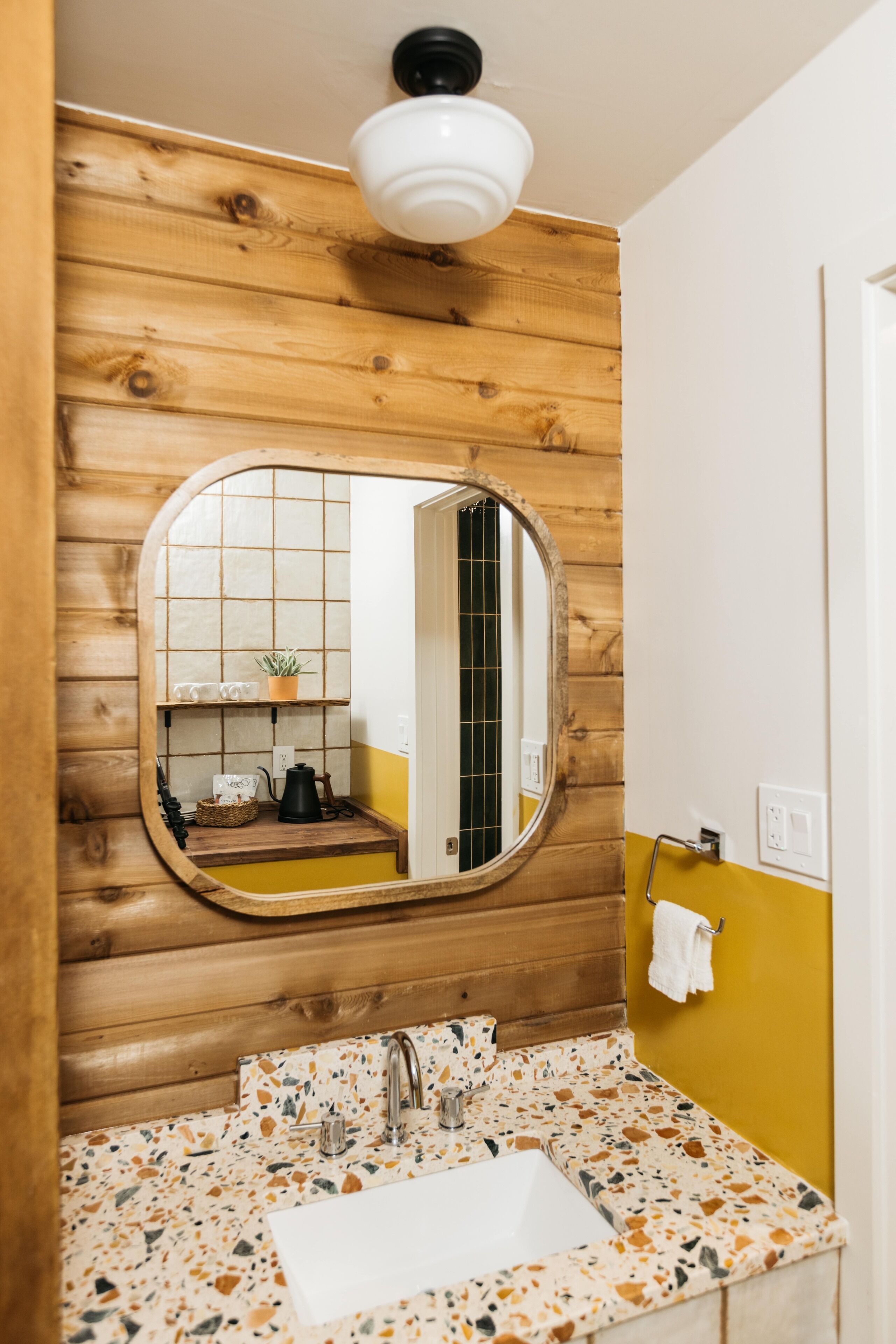 Un baño con pared de madera, un espejo redondo, un lavamanos con encimera de terrazzo y una puerta blanca.