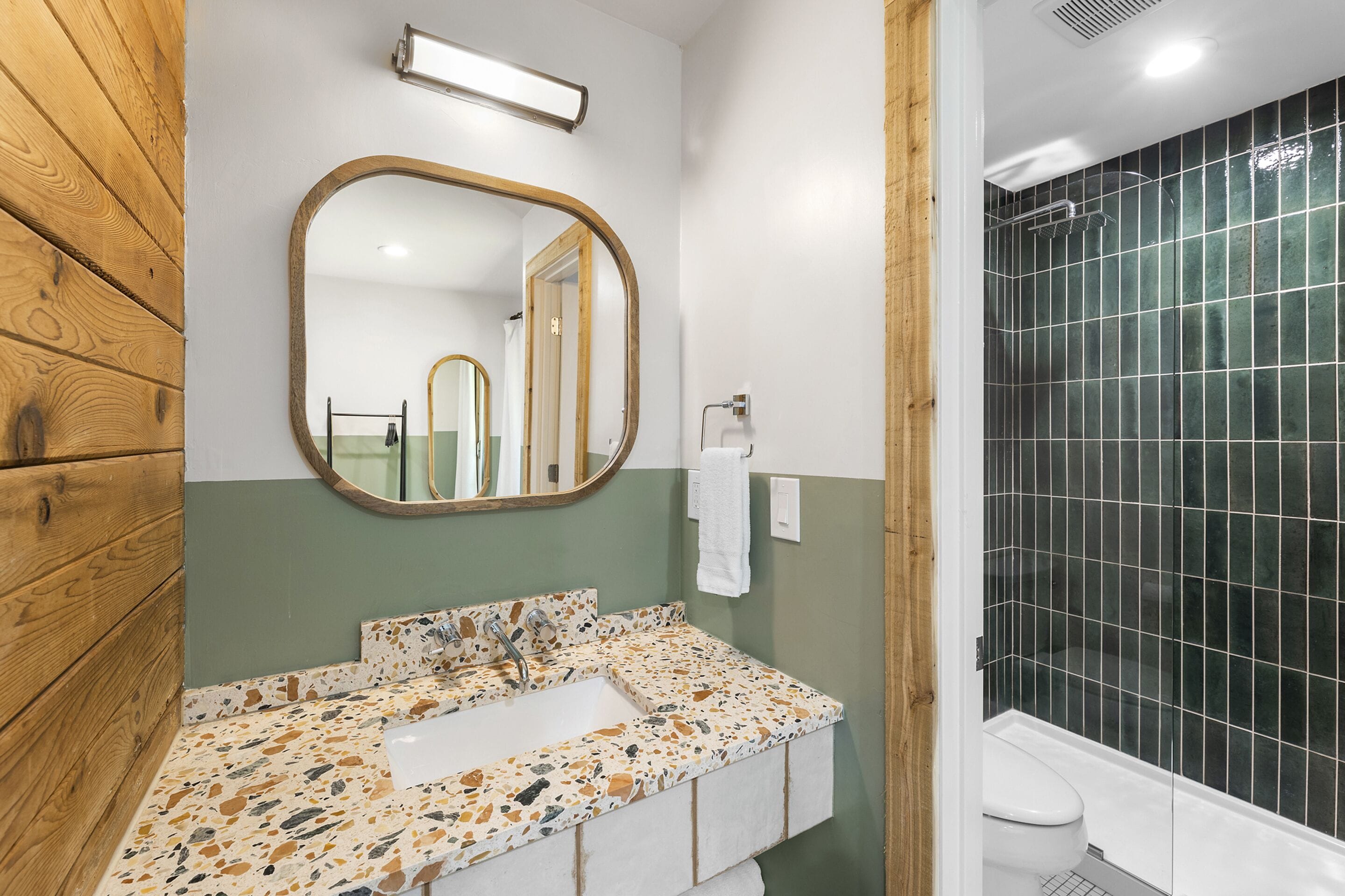 A bathroom with a wooden vanity, a freestanding sink with a speckled countertop, a large mirror, and a shower with dark tiles.