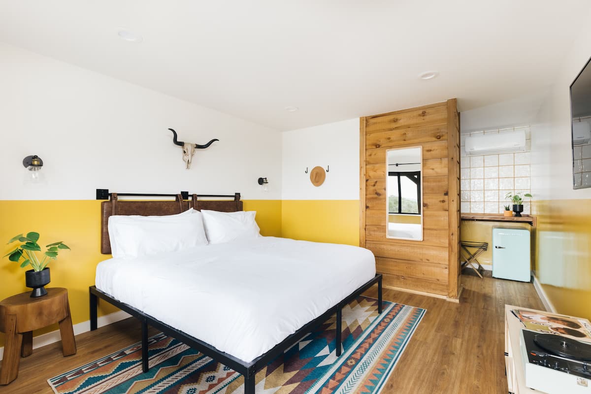 A bedroom with a bed, a wooden headboard, a wall-mounted bull skull, a small stool with a plant, and a kitchen area with a refrigerator and a stove.