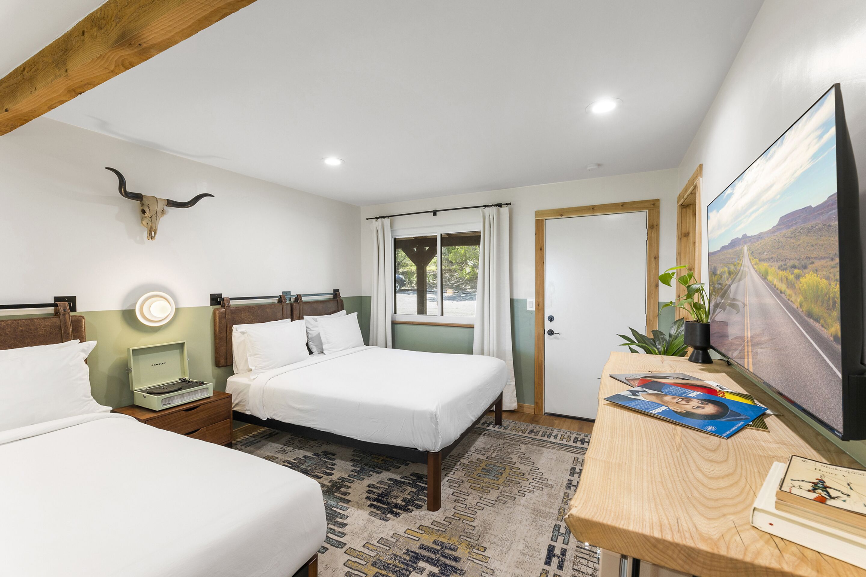 A hotel room with two beds, a desk with a computer and a TV, a bull skull decoration, a window with curtains, and a door.