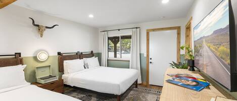 A hotel room with two beds, a desk with a computer and a TV, a bull skull decoration, a window with curtains, and a door.