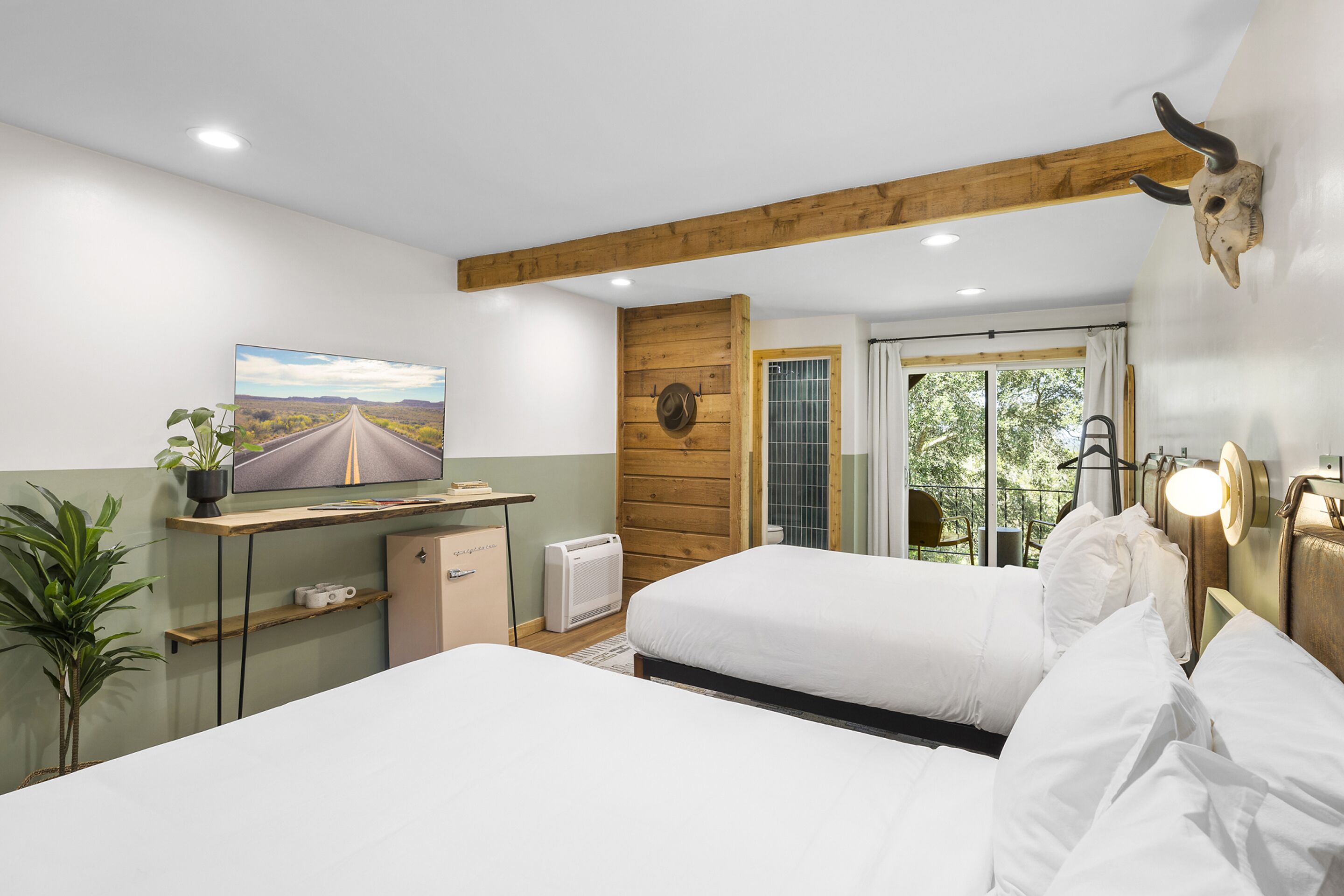 A bedroom with two beds, a TV, a desk, a plant, and a wooden ceiling beam.