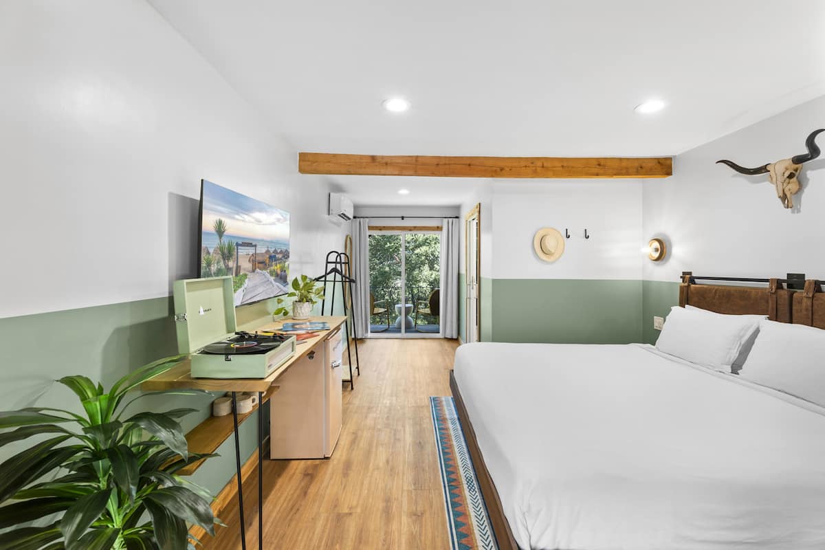 A bedroom with a bed, a desk with a laptop, a potted plant, and a view of the outdoors.