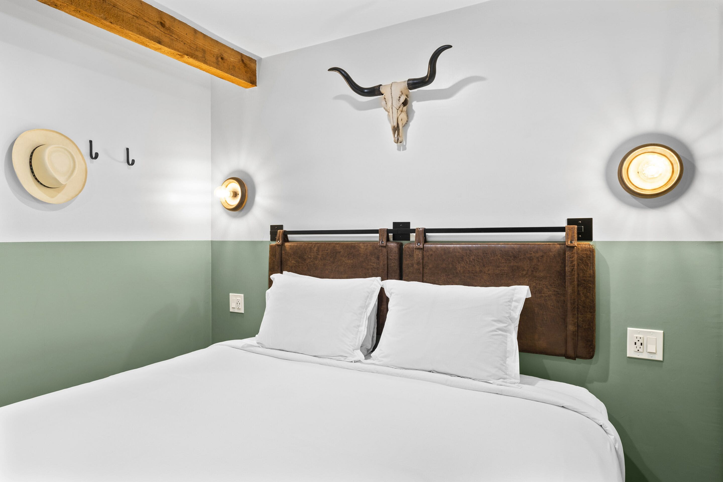 Una cama con cabecera de madera, ropa de cama blanca, una lámpara de pared y un perchero de pared, además de una calavera colgada en la pared.