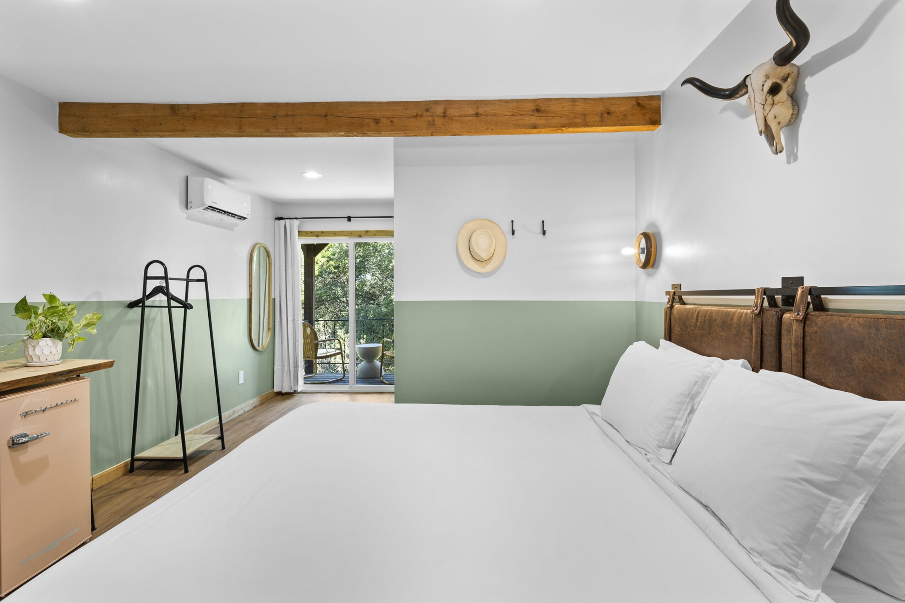 A bedroom with a bed, a wooden ceiling beam, a wall-mounted clock, a hat, and a suitcase.