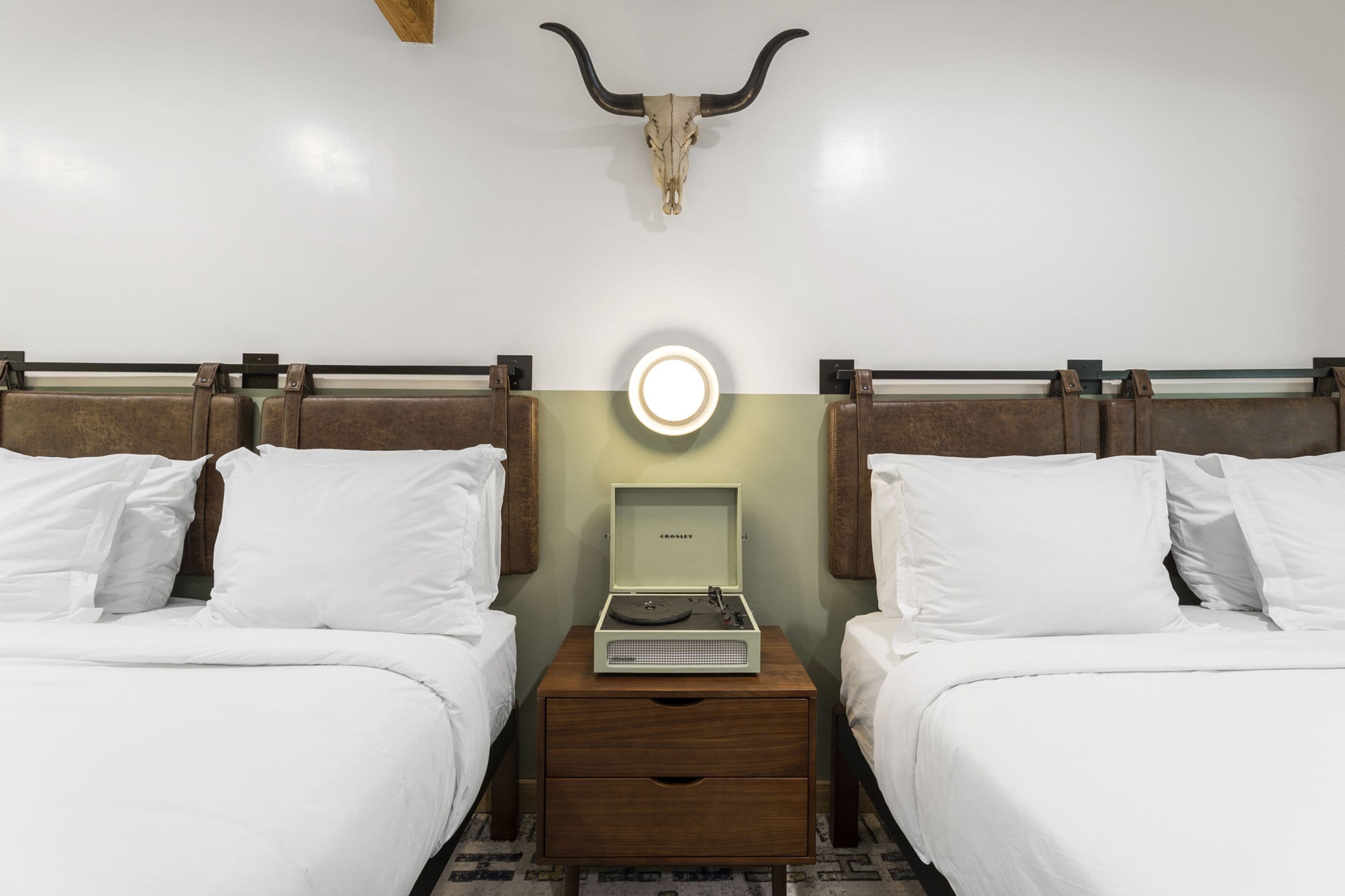 Habitación de hotel con dos camas, una mesita de noche con un tocadiscos, una lámpara de pared y una decoración con cráneo de toro.