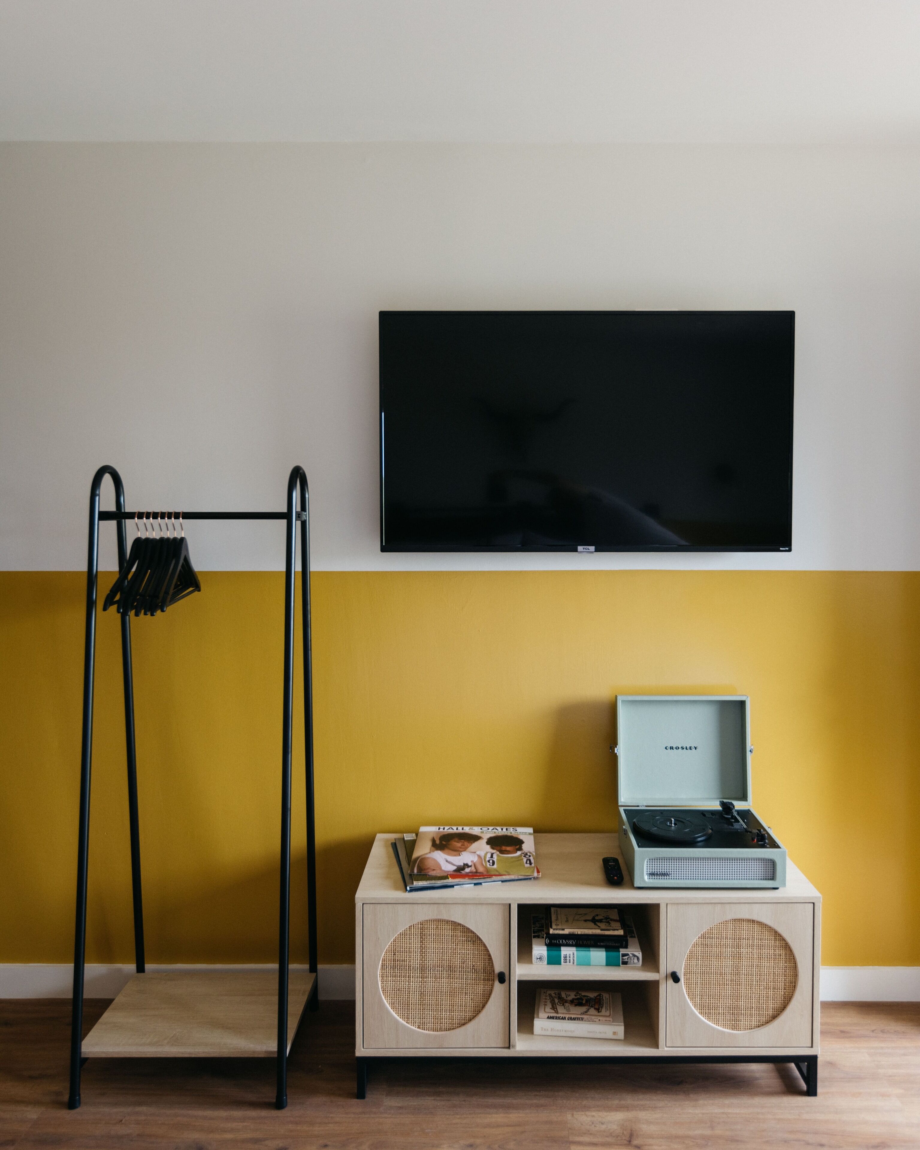 Quarto com televisão na parede, um armário de madeira com toca-discos e um suporte preto para roupas contra uma parede amarela.