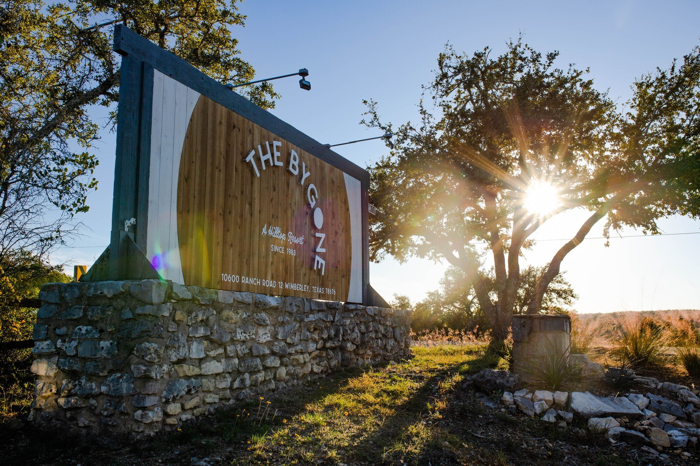 Een houten bord met de tekst 'THE BY GONE' en '1066 RANCH ROAD WIMBERLEY, TEXAS 7878', voor een heldere lucht en een boom waarvan het zonlicht door de bladeren schijnt.