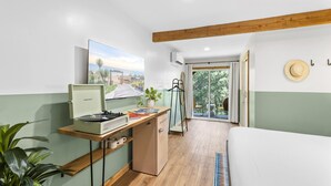 A compact room with a wooden desk, a small refrigerator, a bed, and a large window offering a view of the outdoors.