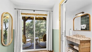 A room with a wooden floor, a wooden bench, a wooden-framed mirror, a bathroom with a wooden vanity, and a sliding glass door leading to an outdoor deck with chairs.