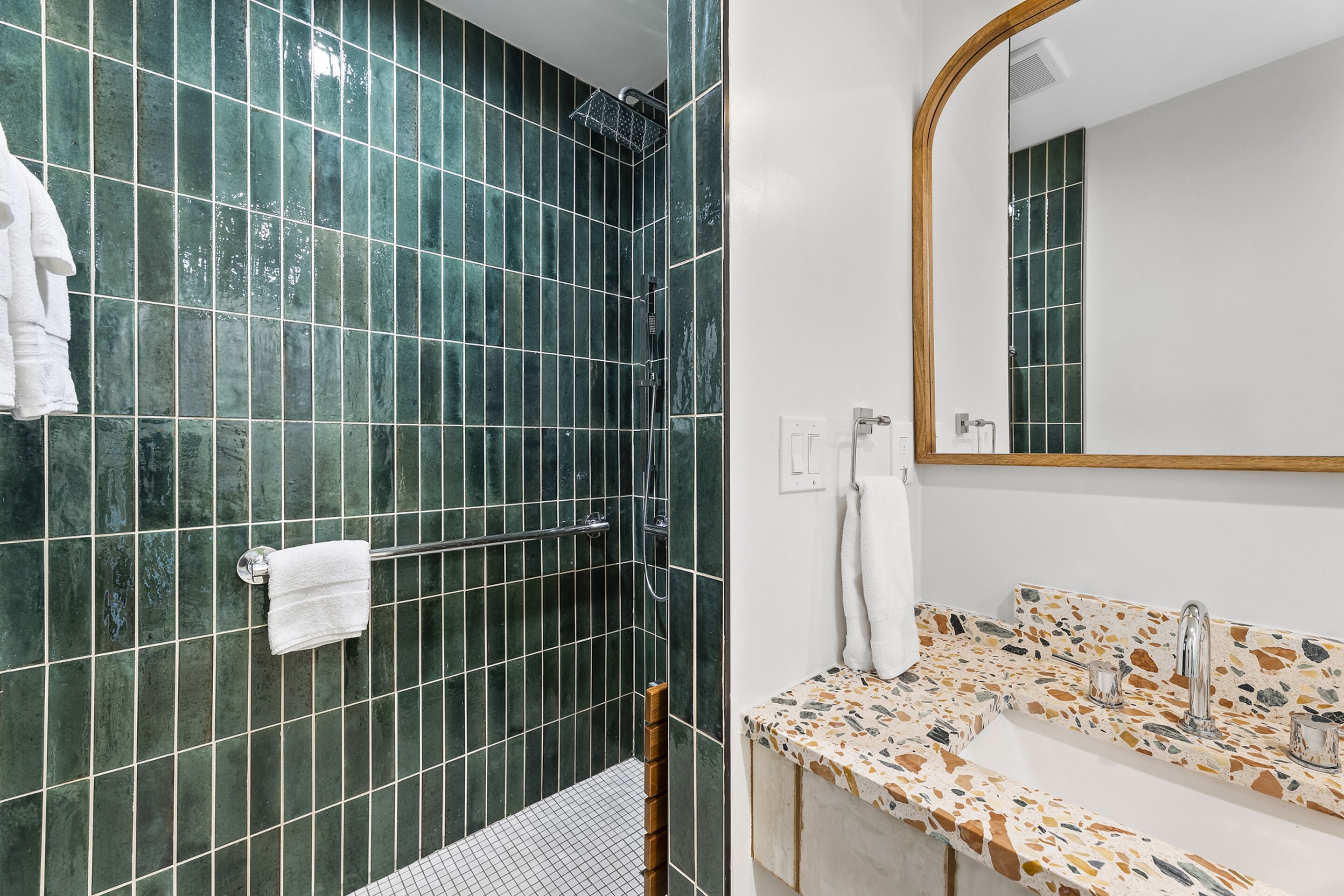 A bathroom with green tiled walls, a shower, a sink with a mosaic countertop, and a mirror with a wooden frame.