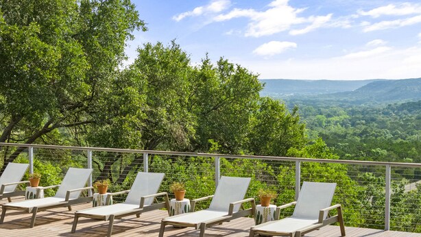 A poolside area with wooden deck and white lounge chairs, surrounded by lush greenery and a scenic view of distant hills.