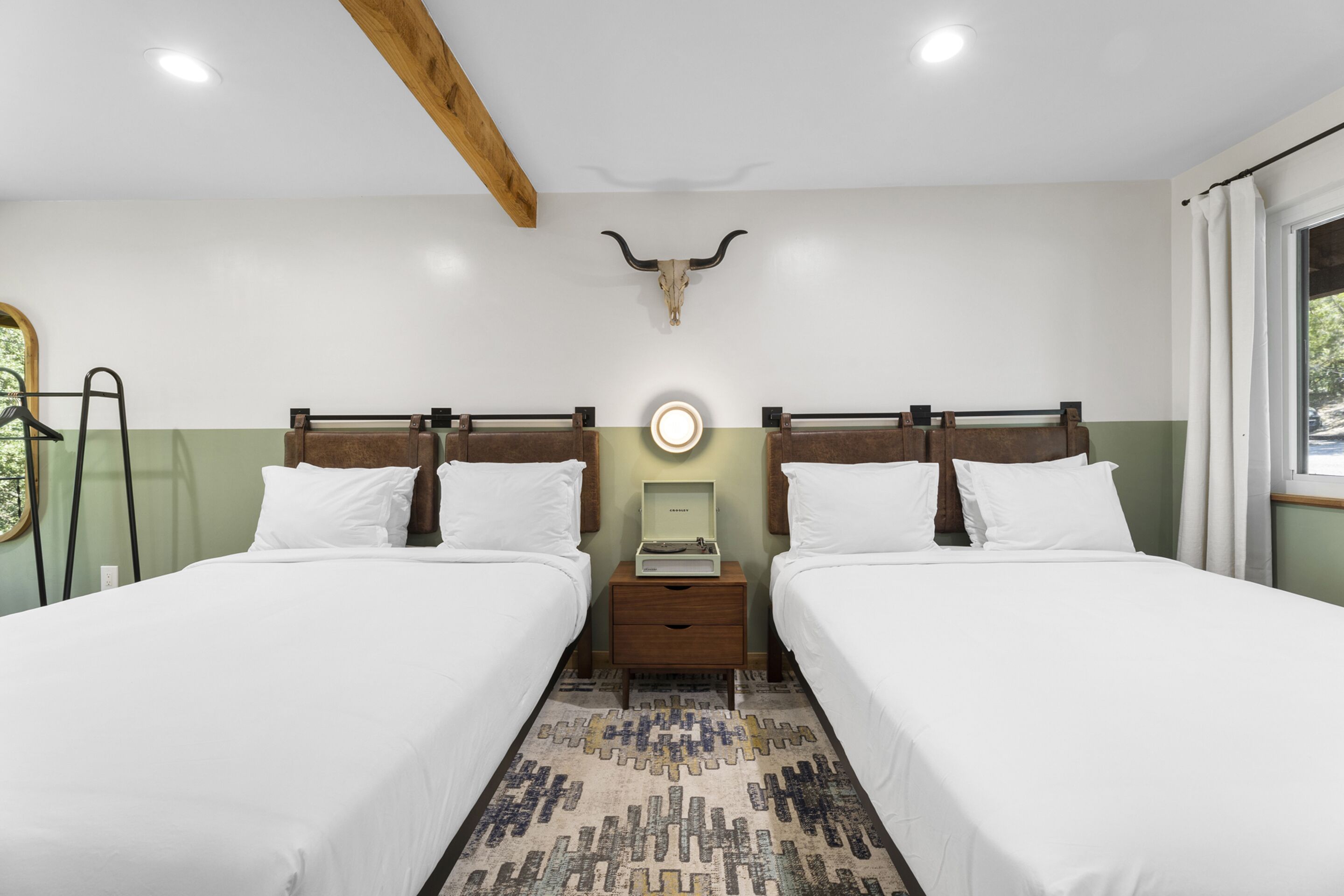 A room with two beds, a wooden ceiling beam, a bull skull decoration, a bedside table with a lamp, and a patterned rug.