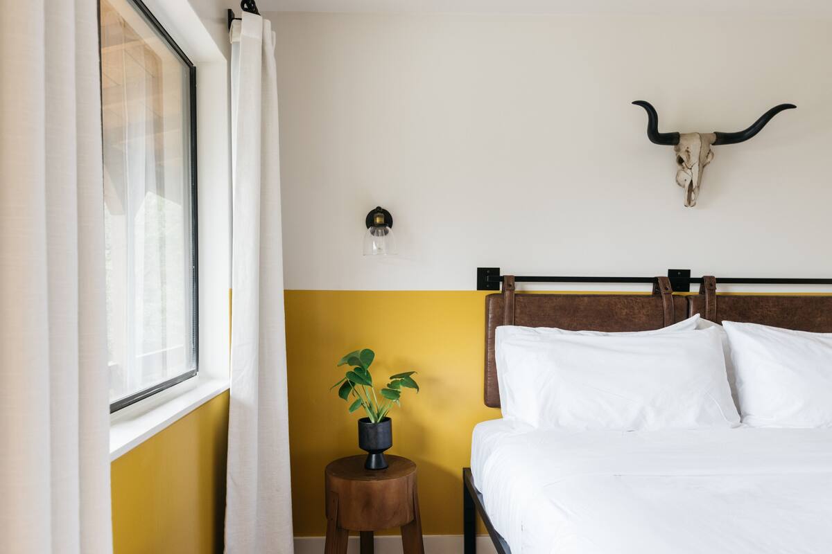 A bedroom with a bed, a window with curtains, a wall-mounted light, a plant, and a cow skull decoration.