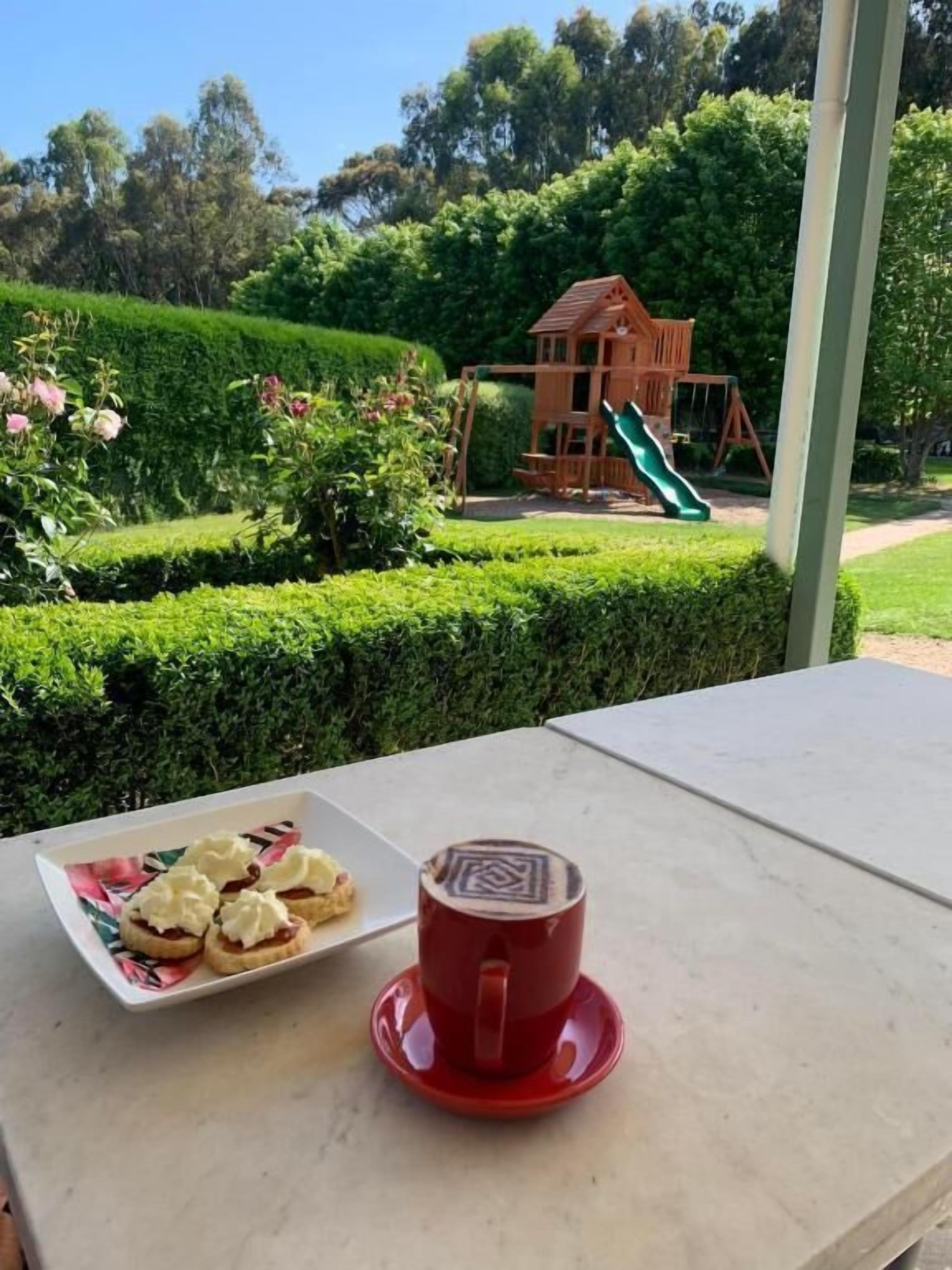 Une table avec une assiette de nourriture et une tasse, le tout placé devant un jardin où se trouve une structure de jeux en bois.
