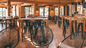 A spacious restaurant with high ceilings, wooden beams, and large windows. Tables and chairs are arranged for dining.