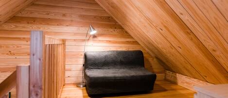 A cozy attic room with wooden walls and a sloped ceiling. A black sofa is positioned against the wall, and a floor lamp is placed nearby.