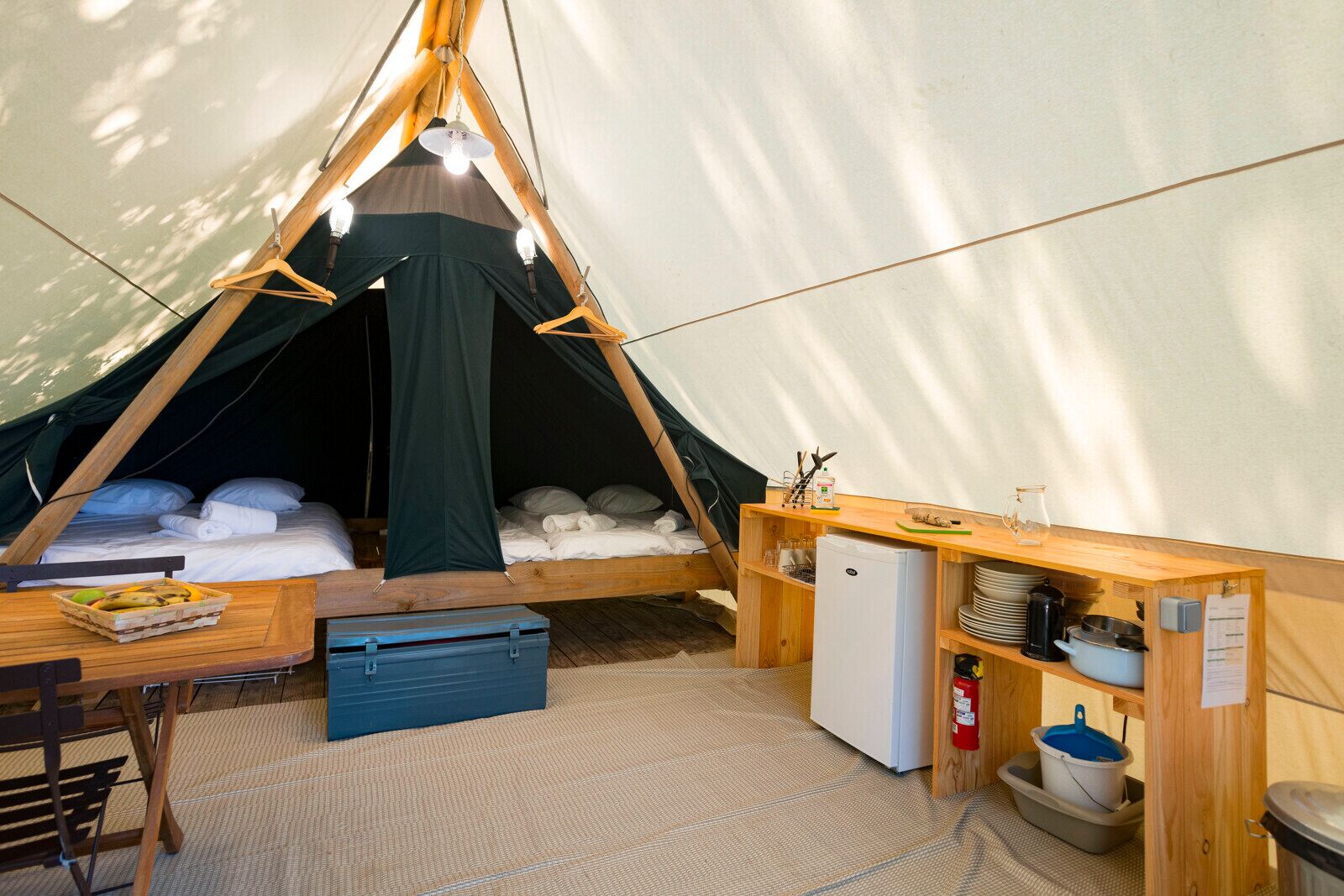 Una carpa con una cama, una mesa con una cesta de fruta, un refrigerador pequeño y un estante con utensilios de cocina.