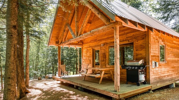A wooden cabin with a porch, a grill, and a view of the forest.