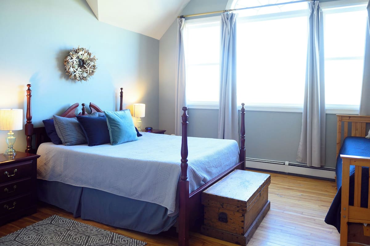 Un dormitorio con cama con dosel, un cofre de madera, un escritorio pequeño y una ventana con cortinas.