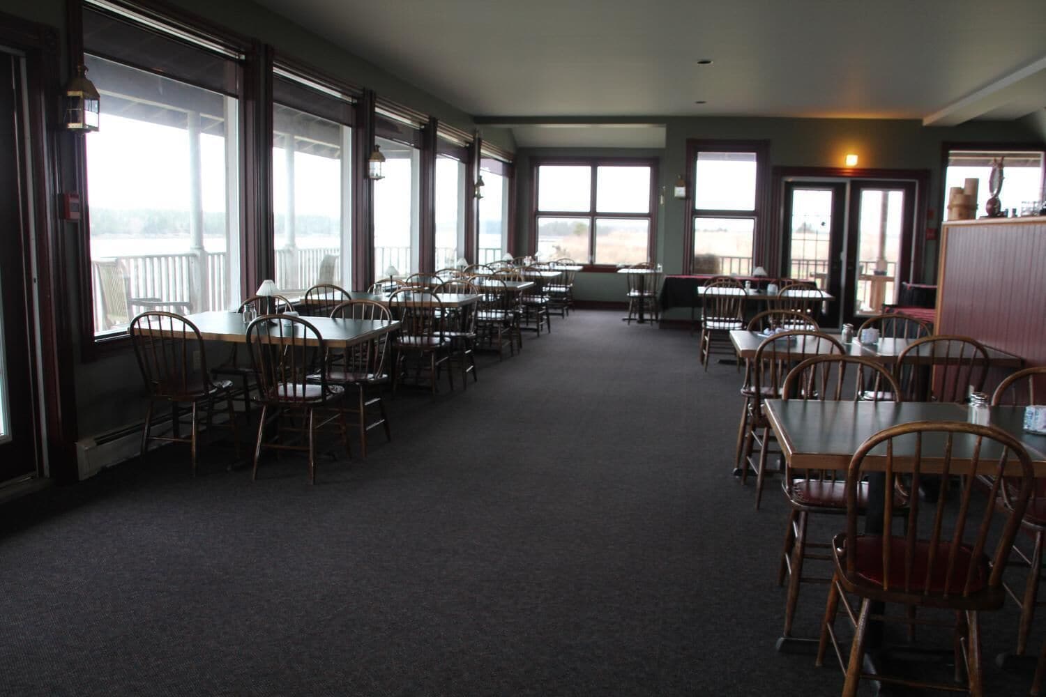 A spacious dining area with multiple tables and chairs, large windows offering views, and a carpeted floor.