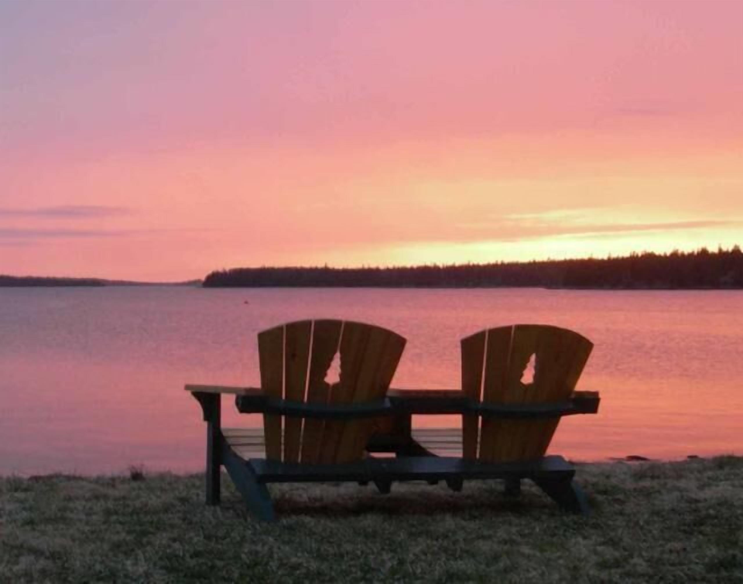 Deux fauteuils Adirondack en bois, placés face à un lac paisible, au coucher du soleil.