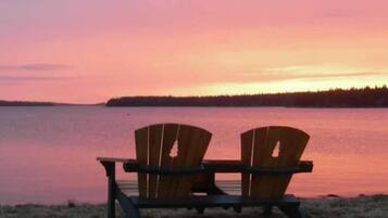 Deux fauteuils Adirondack en bois, placés face à un lac paisible, au coucher du soleil.