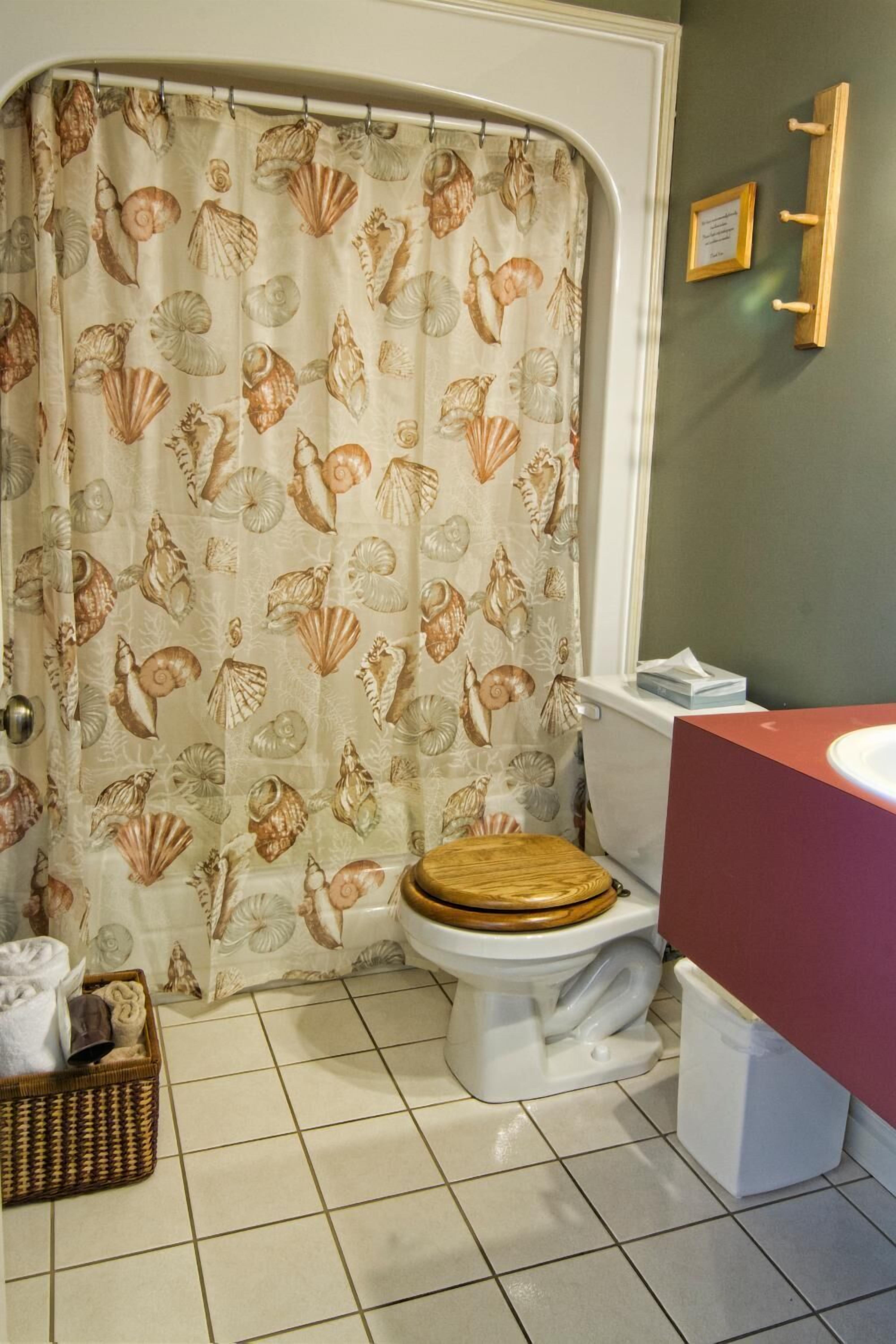 Ein Badezimmer mit Duschvorhang mit Muschelmuster, einer hölzernen Toilettendeckel, einem weißen Waschbecken und einem gefliesten Boden.