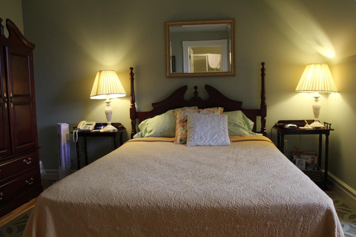 A neatly made bed with a floral pillow, two bedside lamps, a nightstand with a phone, and a mirror on the wall.