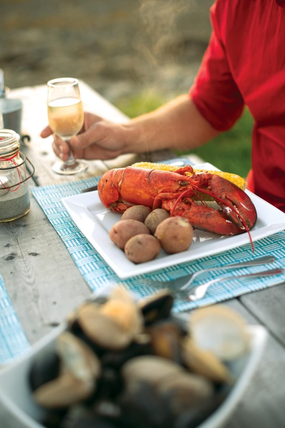 A lobster on a plate with potatoes, a glass of white wine, and a bowl of oysters on a wooden table.